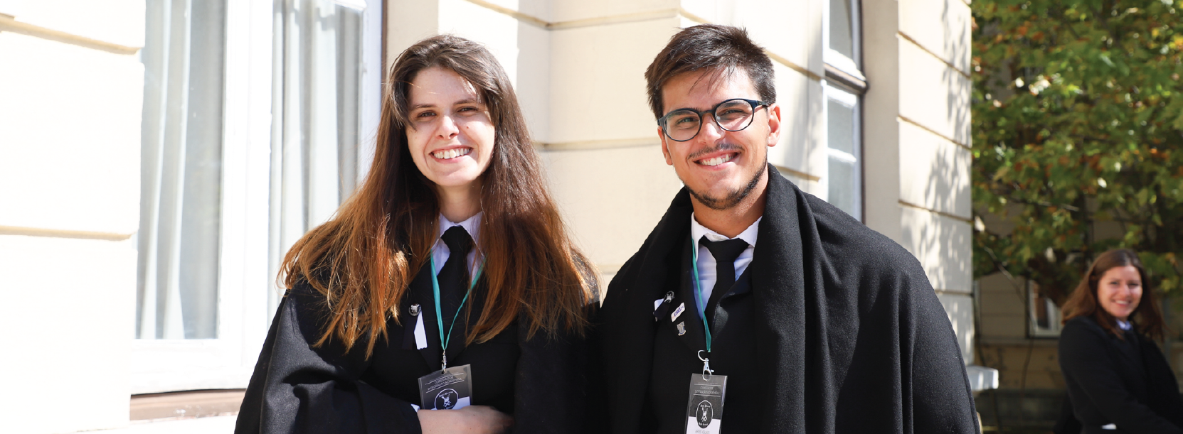 um estudante e uma estudante com traje académico frente à escola